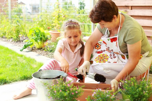 A woman and a young girl are working together in a garden on a sunny day, planting or tending to flowers in a rectangular wooden planter box. The woman is wearing a floral apron and gloves, while the girl, dressed in a light pink top with striped trousers, is focused on the task. In the foreground, there are green plants with small purple flowers in pots, and in the background, a lush garden with a variety of leafy green plants, a wire fence, and a wooden shed or fence panel. The environment appears well-maintained with a neatly mown lawn, and the scene suggests outdoor garden care or landscaping activities near Bermondsey, supporting services related to gardening and patio planting. The natural lighting highlights the vibrant green foliage and colorful flowers, creating a peaceful and productive outdoor space for family gardening or outdoor landscaping enhancements by local Bermondsey landscapers.