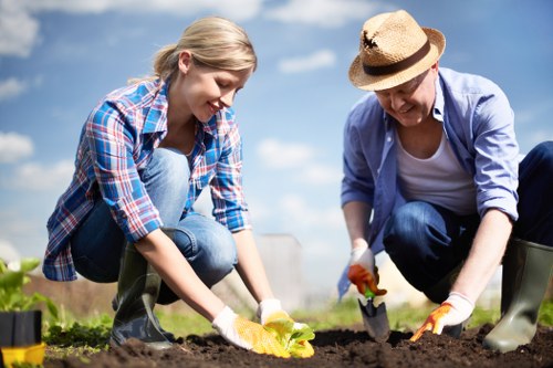 A young woman and an older woman are working together outdoors in a garden, planting or tending to soil. The younger woman, with blonde hair tied back and dressed in a casual plaid shirt and jeans, is carefully placing a yellow flowering plant into the ground. The older woman, wearing a straw hat, light blue shirt, and jeans, is using gardening gloves and a trowel to work the soil around the plant. They are kneeling on dark, freshly turned earth, with small gardening tools and potted plants visible nearby. The background features a bright blue sky with some scattered clouds, indicating a clear, sunny day, in a residential outdoor space that appears well-maintained and suited for gardening activities. The scene exudes a sense of active gardening, emphasizing planting and landscape care, which aligns with gardening and landscape renovation services offered by companies like Landscapers Bermondsey, serving local areas such as Bermondsey and nearby postcodes in London.