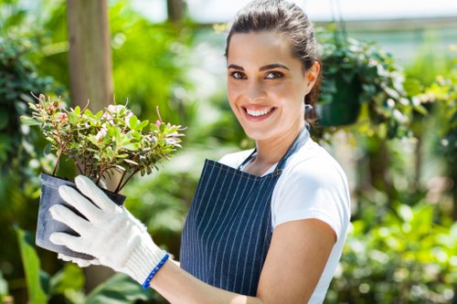 A smiling woman with dark hair tied back, wearing a striped apron and white gardening gloves, stands outdoors in a well-maintained garden in Bermondsey. She holds a potted shrub with green and reddish foliage, positioned in front of a lush backdrop of vibrant green plants, flowering bushes, and trees. The garden features a neat lawn area with dense, healthy grass, bordered by flower beds and a wooden garden fence. Paving stones, possibly part of a pathway or patio, are visible near the edge of the lawn. The lighting suggests a bright, sunny day, with natural light casting soft shadows, highlighting the textures of the soil, foliage, and garden surfaces. The scene reflects typical outdoor gardening activities, indicative of professional landscaping or planting services offered in the Bermondsey area, with emphasis on plant health, garden layout, and maintaining a welcoming outdoor environment.