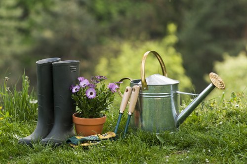 In a domestic garden setting, a pair of black rubber gardening boots are placed on lush green grass alongside a small terracotta pot filled with purple and white flowers. Nearby, there are a variety of garden tools including a pair of pruning shears, a trowel, and a hand fork, all with wooden or plastic handles. A silver metal watering can with a long spout and a round wooden handle is prominently positioned next to the tools. The backdrop features blurred trees and foliage, indicating the garden is outdoor and part of a landscaped yard or back garden. The scene suggests a well-maintained outdoor space suitable for gardening activities, with natural light highlighting the vibrant colours and textures of the plants and garden equipment. This composition aligns with the gardening services offered by Landscapers Bermondsey, focusing on outdoor maintenance and garden care in the Bermondsey area, postcodes SE1 or SE16, with a professional yet natural tone suitable for a service webpage about garden tools and plant care essentials.