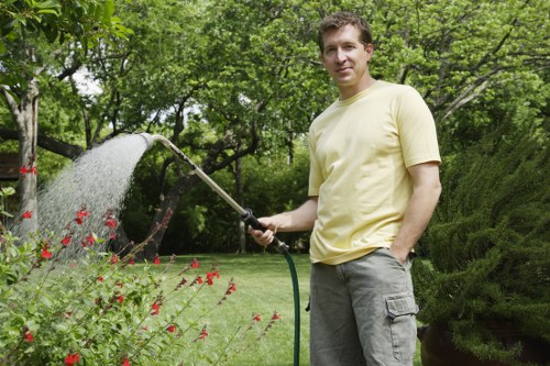 A man in a light yellow T-shirt and grey shorts stands in a well-maintained garden with lush green trees and shrubs in the background. He is holding a garden hose, watering a flower bed that contains red flowering plants and green foliage. The lawn is neatly trimmed, with a combination of grass, paving, and possibly mulch or soil borders visible. The garden appears to be part of a landscaped outdoor space with natural lighting indicating a bright, fair-weather day. The scene reflects outdoor maintenance activity typical of professional gardening services, with a focus on plant care and garden upkeep, relevant to the services offered by Bermondsey-based landscapers.