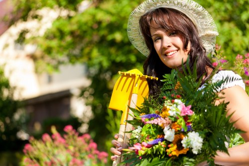 A smiling woman wearing a wide-brimmed straw hat stands in a well-maintained garden, holding a vibrant bouquet of mixed flowers and greenery. Behind her, there is lush green foliage, including shrubs and trees, with some pink flowering plants visible in the background. The garden features a neatly trimmed lawn in the foreground, bordered by a small flower bed containing various colorful plants. The scene suggests a sunny day with natural light illuminating the garden and highlighting the textures of the grass, plants, and the woman's floral arrangement. This outdoor space is typical of residential gardens in Bermondsey, showcasing well-kept landscaping and planting areas that could be part of a professional gardening service such as those offered by Landscapers Bermondsey, fitting within their gardening and planting solutions in the local area.