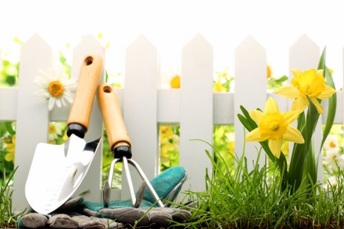 The image depicts a section of a garden with a white picket fence in the background, adorned with vibrant yellow daffodils and white daisies in full bloom. In the foreground, there is a small bed of lush green grass with a healthy, dense texture, alongside a patch of freshly turned dark soil. Leaning against the fence are gardening tools, including two small hand shovels with wooden handles and a hand rake, suggesting recent or ongoing gardening work. The scene is well-lit with natural sunlight, indicating a bright and clear day, which highlights the lively colours and fresh greenery. This outdoor space exemplifies a well-maintained garden suitable for appealing landscaping and lawn care services offered by Bermondsey-based garden specialists. The overall setting suggests a tidy, inviting yard with a simple yet charming aesthetic, appropriate for family gardening or outdoor relaxation, and aligns with professional landscape gardening in the Bermondsey area.