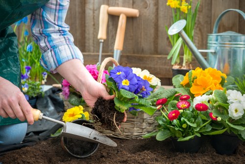 A gardener is in a backyard garden, preparing to plant flowering plants in a garden bed with dark, moist soil. The gardener's hand is inserting a potted plant with vibrant purple and yellow flowers into the ground, while a trowel rests in the soil nearby. In the background, there are additional potted plants with colorful blooms, including yellow, pink, and red flowers, along with gardening tools, a watering can, and a wooden fence. The garden area features a mix of soil, potted plants, and garden equipment, indicating active planting and outdoor maintenance typical of residential outdoor spaces in Bermondsey. Natural daylight illuminates the scene, highlighting the rich textures of the soil and the vivid colours of the flowers, supporting services related to gardening, landscaping, and outdoor plant care.