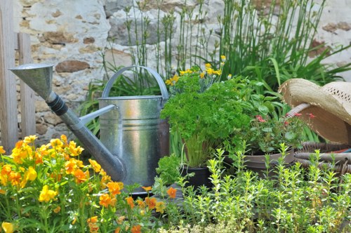 The image depicts a small garden space with a variety of plants and garden elements arranged in a natural and casual manner. In the foreground, there is a dense bed of vibrant orange and yellow flowering plants, possibly marigolds, providing bright splashes of colour. Behind this flower bed, lush green foliage with small, herb-like leaves is visible, adding texture and fullness to the garden. To the right, there is a rustic, wide-brimmed straw hat resting on a dark plant pot, suggesting outdoor leisure or gardening activity. A weathered, metal watering can with a rounded handle is prominently positioned in the garden, indicative of routine maintenance. Tall, green grasses or reeds grow along the back, their upright blades adding height to the scene. The background features a light-colored brick or stone wall that encloses the garden space, giving an enclosed, cozy outdoor environment. The overall setting appears to be a well-maintained but relaxed garden area suitable for outdoor gardening and leisure activities near Bermondsey, with natural daylight illuminating the scene and subtle signs of ongoing care by gardening professionals such as Landscapers Bermondsey.