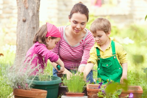 A woman and two young children engage in gardening outdoors in a backyard with lush greenery, including a large tree trunk in the background. The woman, wearing a pink and white striped top, is smiling as she helps the children plant or tend to small plants or flowers in various terracotta and plastic pots arranged on a garden table. One child, dressed in a pink hoodie, is focused on handling a small gardening tool, while the other, wearing a yellow shirt with green overalls, observes nearby. The garden features a well-maintained lawn with visible grass patches, flower beds, and a few shrubs, creating a vibrant and inviting outdoor space typical of residential gardens in Bermondsey. The scene is illuminated by natural, possibly late afternoon sunlight, highlighting the bright colours of the children’s clothing and the greenery. This image relates to professional gardening services offered by Landscapers Bermondsey, emphasizing outdoor planting, garden maintenance, and family-friendly landscape enhancement in the local area.